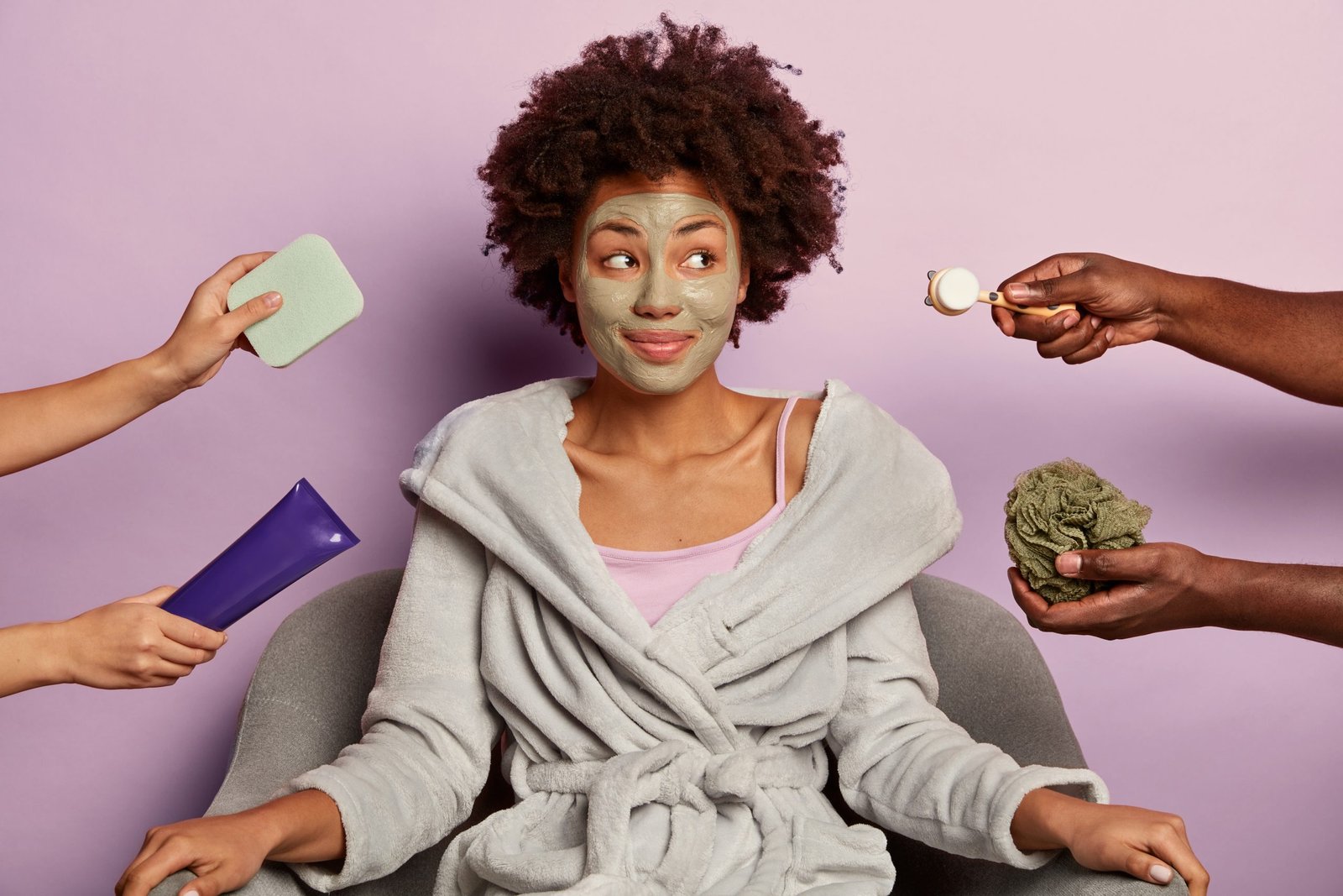 Relaxed curly haired woman wears clay mask on face, cares about facial skin, gets various cosmetic treatments after taking bath, looks aside, sits in comfortable armchair, wears domestic clothes
